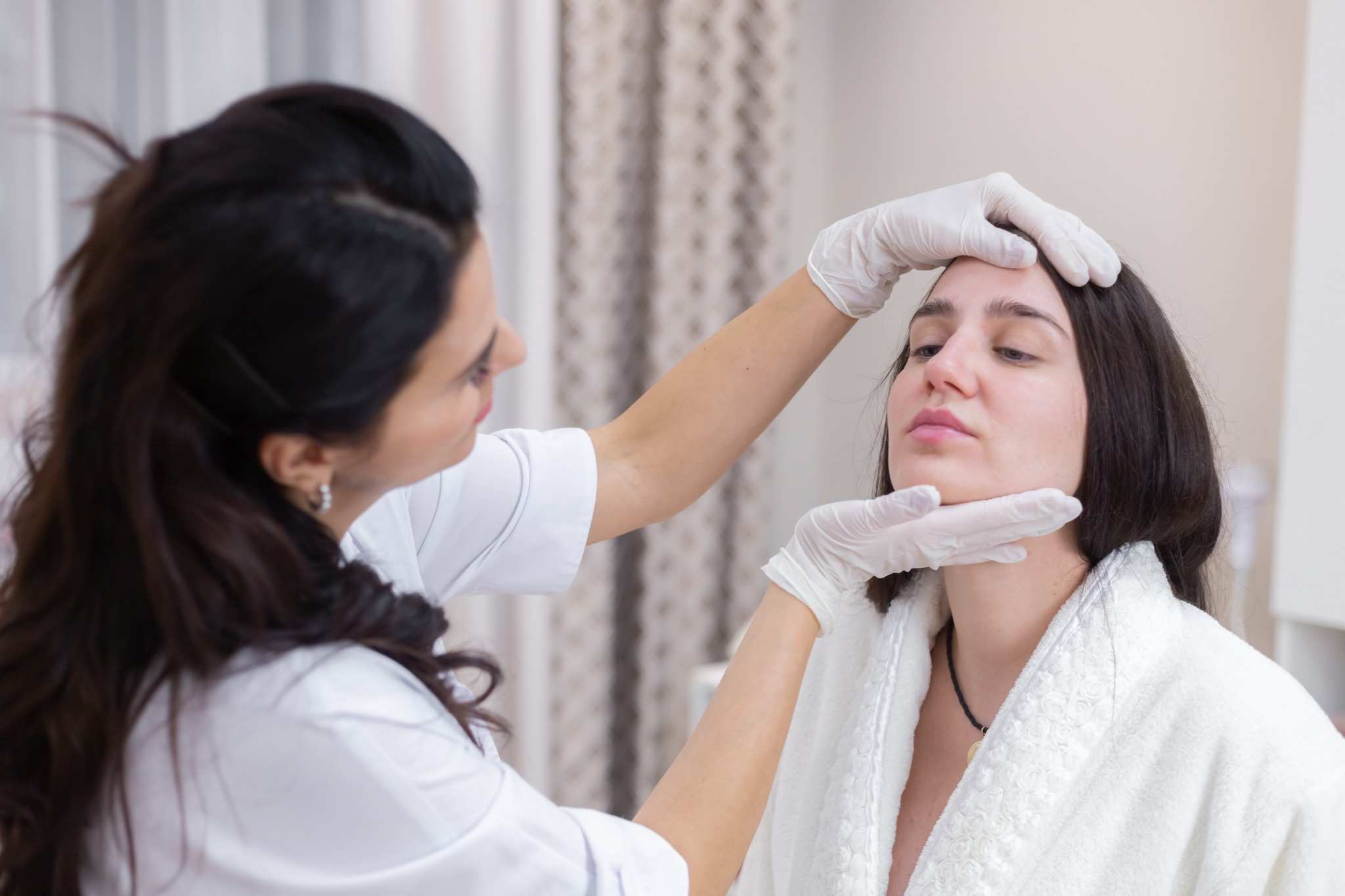 Aesthetic practitioner wearing gloves examining a woman’s face during a cosmetic consultation in a clinic setting.
